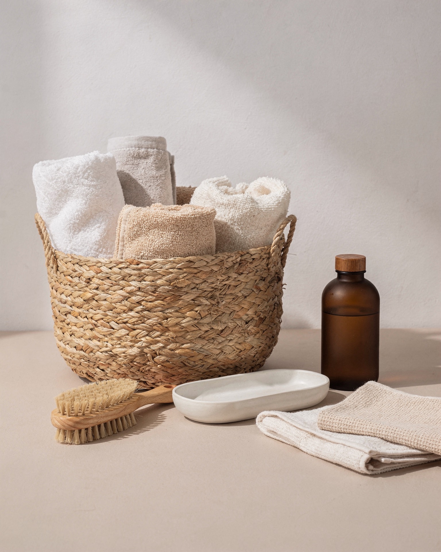 A woven basket of folded towels with a wooden brush, ceramic tray, and amber bottle arranged as a simple home reset station.